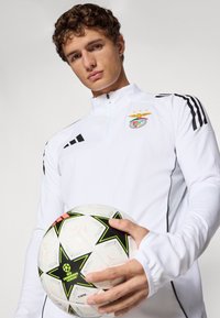 Ung mann i hvit sportsjakke med Benfica-logo holder en UEFA Champions League fotball mot en ensfarget bakgrunn.