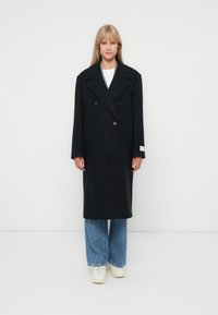 Abrigo negro de lana de doble botonadura con solapas anchas y dos botones, combinado con jeans anchos de lavado claro y zapatillas blancas.