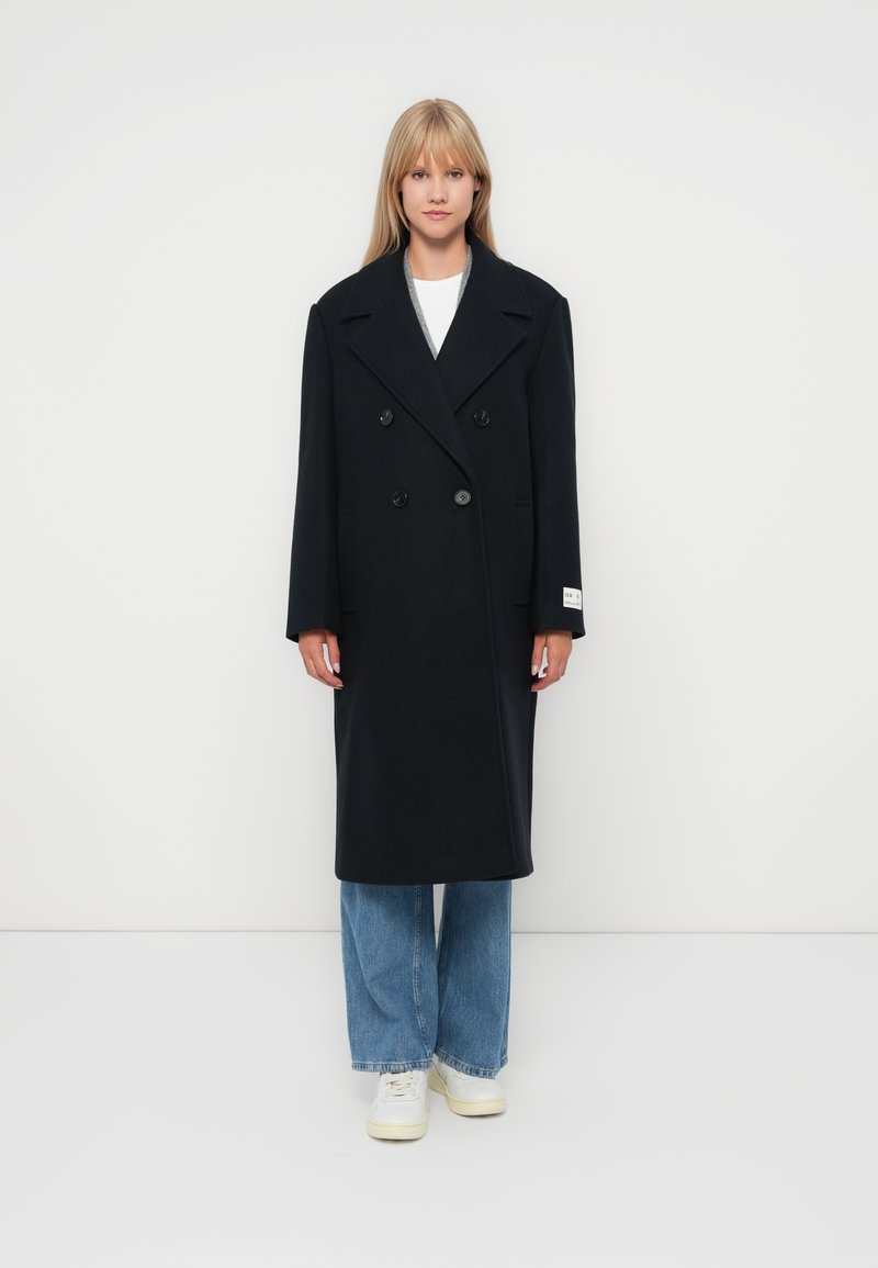 Abrigo negro de lana de doble botonadura con solapas anchas y dos botones, combinado con jeans anchos de lavado claro y zapatillas blancas.