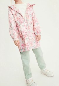 Veste de pluie florale rose clair avec capuche, fermeture éclair à l'avant et poignets élastiques ; associée à un pantalon vert menthe et des baskets blanches.