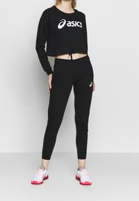 Zwarte, cropped sweatshirt met wit "asics" logo, gecombineerd met zwarte, aansluitende joggers met een accentlogo. Model draagt kleurrijke sportschoenen.