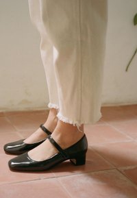 Scarpe Mary Jane in vernice nera con tacco basso a blocco, dotate di punta quadrata e una sola fascetta trasversale. Indossate con pantaloni crema sfrangiati.
