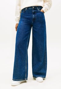 Niebieskie jeansy o szerokich nogawkach z wysokim stanem, wyposażone w przednie kieszenie i jasne przeszycia. Noszone z białymi trampkami.