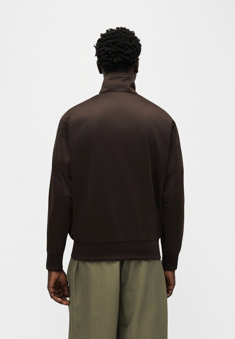 Uomo con capelli ricci scuri che indossa una giacca marrone scuro con collo alto e pantaloni verde oliva, in piedi di spalle contro uno sfondo semplice.