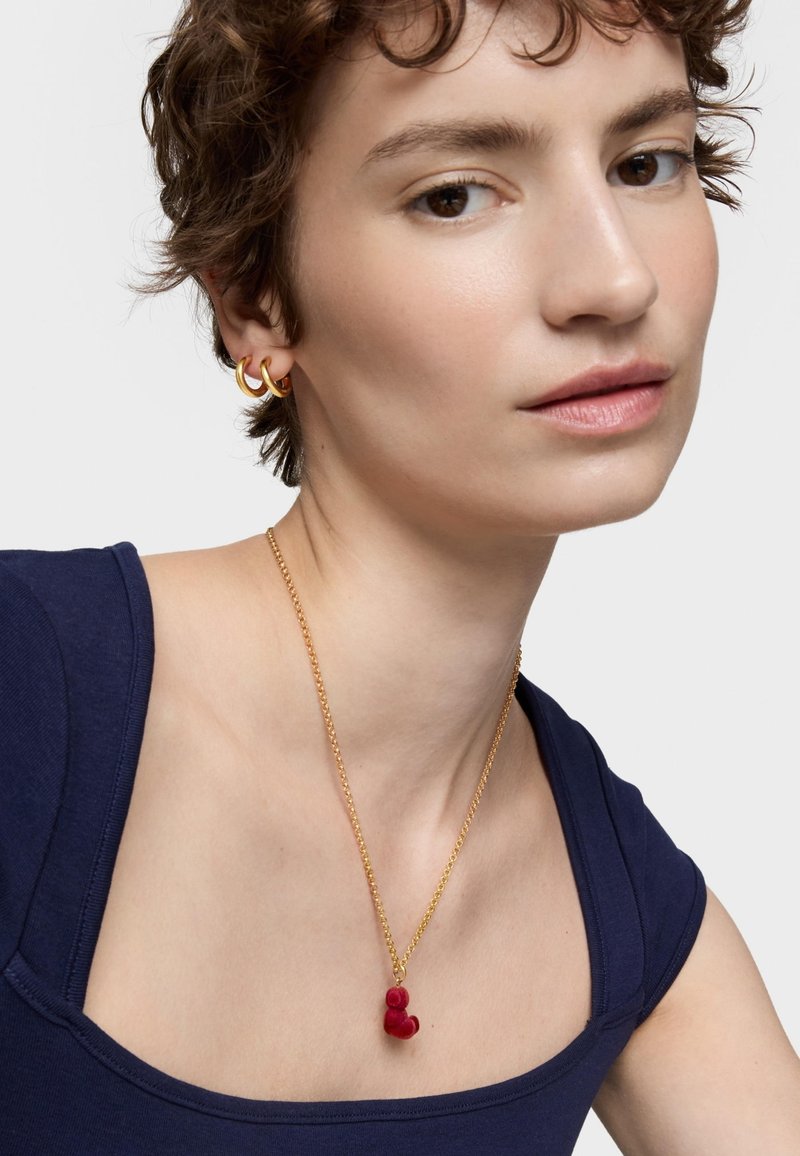 Collar de cadena de oro con un colgante de terciopelo rojo, llevado con aretes de aro dorados, sobre una camisa azul marino. Fondo neutro.