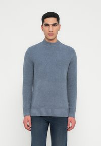 Fiatal férfi előrefelé néz, világoskék kötött kereknyakú pulcsit és sötétkék farmert visel, háttérben egy sima fehér fal.