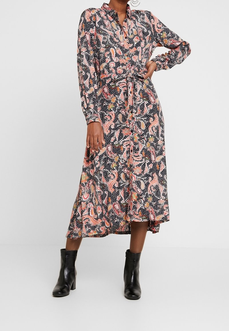 Femme portant une robe noire à manches longues boutonnée avec un motif cachemire rose et des pois blancs, associée à des bottines noires à talons.
