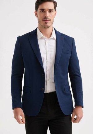 Homme portant un blazer bleu marine, une chemise blanche et un pantalon noir, debout devant un fond clair et uni.