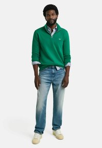 Grøn sweater med kvart-zip krave over en lyseblå skjorte, parret med falmede blå jeans og hvide sneakers. Enkel design og afslappet pasform.