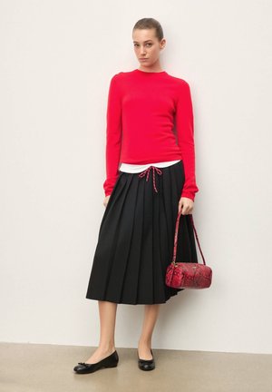 Kobieta ubrana w czerwony sweter, czarną plisowaną spódnicę midi, czarne baleriny, trzymająca czerwoną torebkę z motywem skóry węża, stoi na tle białej ściany.