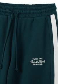 Pantalones de chándal de tiro alto en verde oscuro con cordón, con una franja lateral blanca y texto bordado en blanco "París 1996 Rue de Rivoli Sport Club" en el muslo izquierdo.