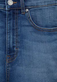 Tela de denim azul índigo que presenta un bolsillo frontal con costuras amarillas, un cierre de botón metálico y una textura suave con un leve desgaste.