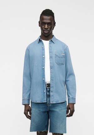 Mann trägt hellblaues Jeanshemd über weißem T-Shirt und passende Jeansshorts mit rohem Saum, posiert vor schlichter weißer Wand.