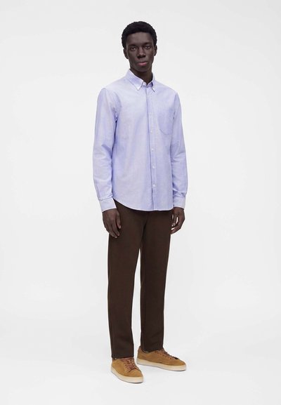 Homme debout portant une chemise bleu clair boutonnée, un pantalon marron foncé et des baskets en daim beige sur fond blanc uni.