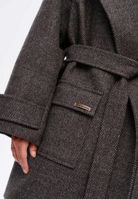 Bruine tweedjas met een riem, brede mouwen en een patch pocket met een "blue shadow" logo label, die een gestructureerd geweven patroon laat zien.