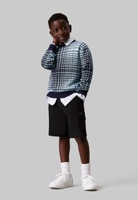 Maglione a quadri blu e blu navy, pantaloni cargo neri, camicia bianca con colletto, sneakers bianche e calzini bianchi. Outfit casual a strati per bambini.