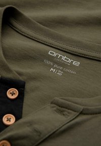 Olivgrön bomullsskjorta med krage, svart knäppning, bruna knappar och tryckt etikett med texten "ombre cotton line 100% ren bomull M/L 48".