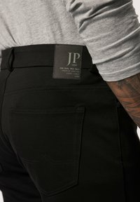 Pantalon noir en tissu texturé, avec une poche arrière dotée d'un motif de couture décoratif et une patte en cuir estampillée au niveau de la taille.