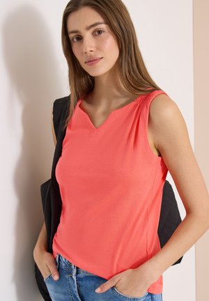 Jonge vrouw met lang bruin haar, draagt een koraalkleurige mouwloze top en blauwe spijkerbroek, leunend tegen een lichtgekleurde muur met handen in de zakken.