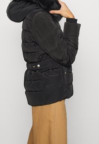 Černá péřová bunda s kapucí, zdobená límcem z umělé kožešiny, prošívaným designem, bočními kapsami a akcenty na kovové cvoky.
