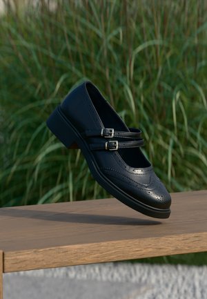 Sort læder Mary Jane-sko med dobbelte spænder og brogue-detaljer, der svæver over en træbænk med grøn græsbaggrund.