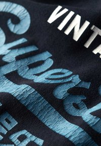 Zwarte stof met verouderde blauwe tekst met een gestructureerde afwerking, gedeeltelijk verborgen witte letters, die een vintage ontwerp benadrukken.