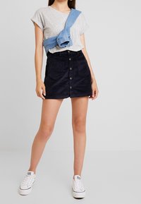 Kvinna bär en grå t-shirt, en knappad marinblå kordkragsminikjol, vita sneakers och en ljusblå jeansjacka över bröstet.