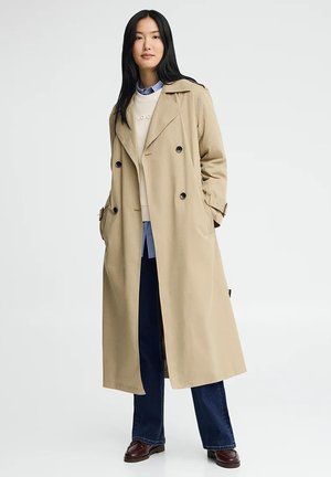 Kvinde med langt sort hår iført beige dobbeltradet trenchcoat, blå jeans, hvid sweater og brune loafers, stående med hænderne i lommerne.
