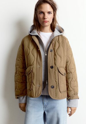 Jeune femme portant une veste matelassée beige par-dessus un sweat à capuche gris zippé et un jean bleu clair, debout devant un fond blanc uni.