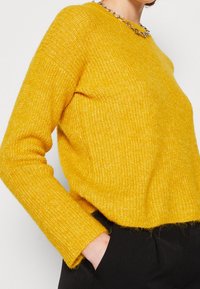 Gul stickad tröja med en texturerad yta, rundad halsringning och långa ärmar. Accentuerad med ett silverkedjehalsband. Svarta byxor synliga.