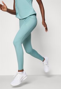 Leggings de sport teal clair avec une coupe ajustée, texture lisse et accents perforés sur les côtés, associés à des baskets blanches.