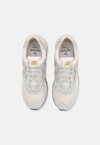 Tenisky New Balance v světlých šedých a béžových barvách, hladký povrch, tkaničky, zlaté akcenty a klasický design.