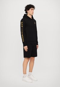 Black hoodie with front kangaroo pocket, hood, and yellow EA7 branding on the sleeve. Paired with black shorts and white sneakers.