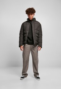 Sort puffjakke med høj krave, forlommer og en glat tekstur. Kombineret med en sort hoodie og beige bukser, afsluttet med sorte sneakers.