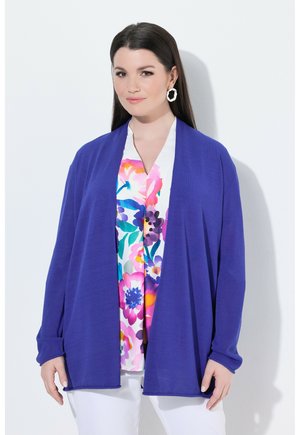 Femme aux longs cheveux foncés porte un pantalon blanc, un chemisier fleuri coloré, un cardigan bleu roi ouvert sur le devant et des boucles d’oreilles blanches en forme d’anneau.