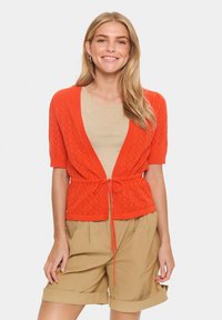 Korte mouwen oranje cardigan met een strik in de taille, met een gebreid patroon. Draag het over een beige top en combineer het met beige shorts.
