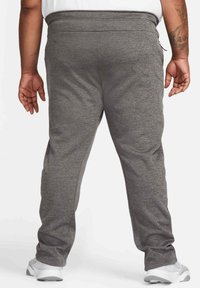 Homme portant un pantalon de survêtement gris et des baskets blanches, photographié de dos sur un fond uni.