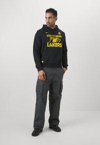 Hoodie preto com o logótipo amarelo dos "Los Angeles Lakers", emparelhado com calças cargo cinzentas com bolsos laterais e sapatilhas pretas.