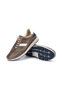Zapatilla de cuero marrón con superficie texturizada, acentos blancos y detalles cosidos. Presenta una suela de goma con un patrón azul y un diseño bicolor.