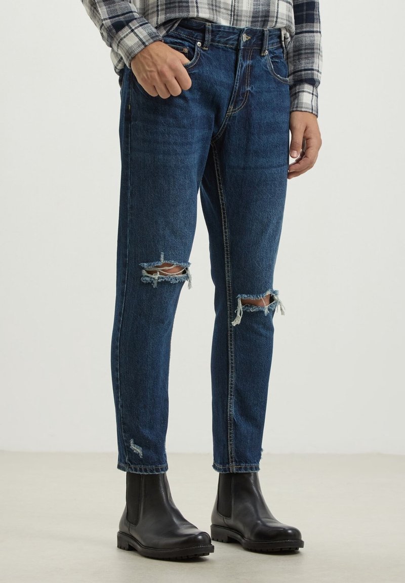 Man wearing blue ripped jeans, black leather boots, and a gray plaid shirt standing against a plain background.