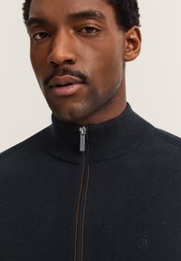 Maglione nero con zip, colletto alto, texture a costine e logo discreto sul petto. Dettaglio della zip evidente sulla parte anteriore.