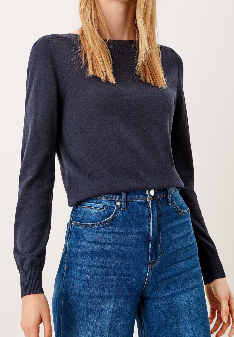 Pull-over bleu marine à manches longues avec encolure côtelée, texture douce et poignets ajustés, associé à un jean bleu taille haute avec poches avant.