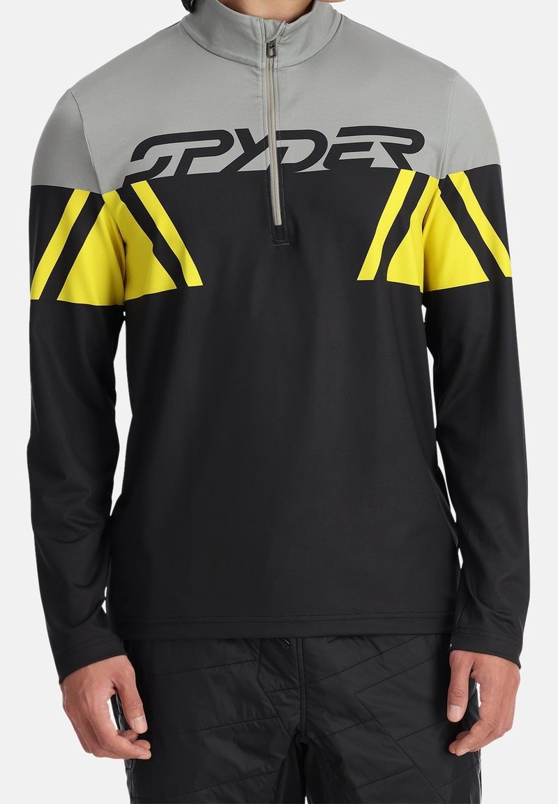 T-shirt de sport à manches longues noir, gris et jaune avec fermeture éclair à l'avant et logo audacieux "SPYDER" sur la poitrine, porté par une personne.