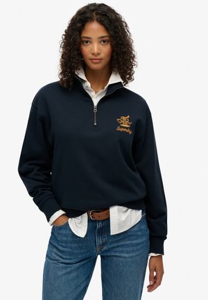 Sudadera navy con cierre y puños de canalé, que presenta un logo bordado en dorado. Llevada sobre una camiseta blanca y combinada con jeans azules.