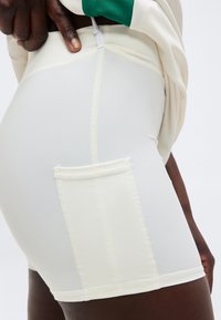 Κοντινό πλάνο ατόμου που φοράει λευκά εφαρμοστά σορτς με πλαϊνή τσέπη και πράσινο-άσπρο τοπ, με το χέρι να ρυθμίζει το φερμουάρ στη μέση.