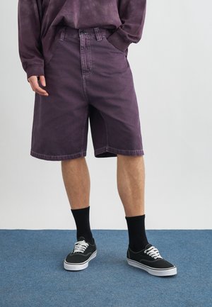 Άτομο που φοράει μοβ τζιν σορτς, μακρυμάνικο πουκάμισο που ταιριάζει, μαύρες κάλτσες και μαύρα sneakers με κορδόνια, στέκεται σε μπλε χαλί.