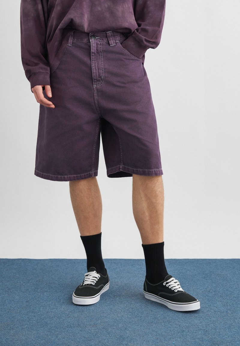 Άτομο που φοράει μοβ τζιν σορτς, μακρυμάνικο πουκάμισο που ταιριάζει, μαύρες κάλτσες και μαύρα sneakers με κορδόνια, στέκεται σε μπλε χαλί.