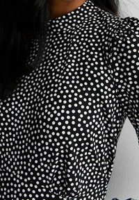 Svart klänning i tyg med vita prickar, med hög hals och puffiga ärmar. Tyget ser slätt och lätt ut.