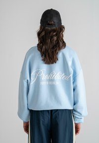 Helle blaue übergroße Sweatshirt mit weißem Schriftzug "Prohibited 10119 BERLIN" auf der Rückseite. Kombiniert mit dunkelblauen Jogginghosen.
