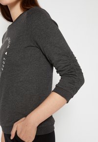 Dunkelgrauer Sweatshirt mit langen Ärmeln, rundem Ausschnitt, gerippten Bündchen und einem bedruckten Design auf der Vorderseite. Glatte Stofftextur.